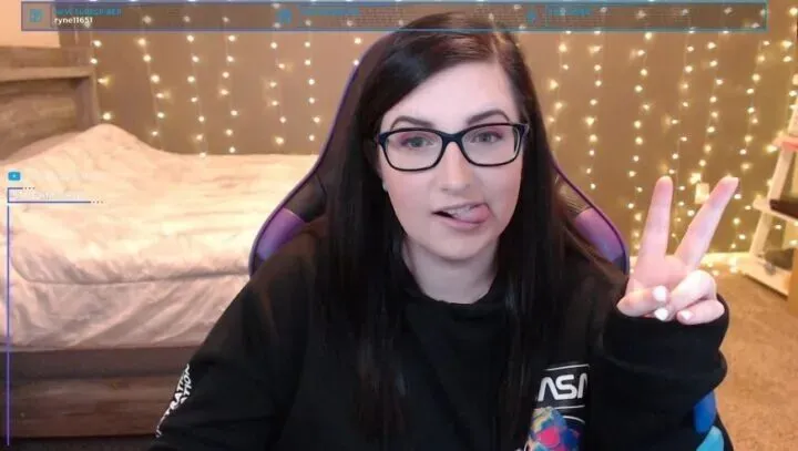 ShelbyRenae screengrab - girl with glasses is shown holding up a peace sign with her tongue sticking out, wearing a black long sleeve shirt and she has brown hair.